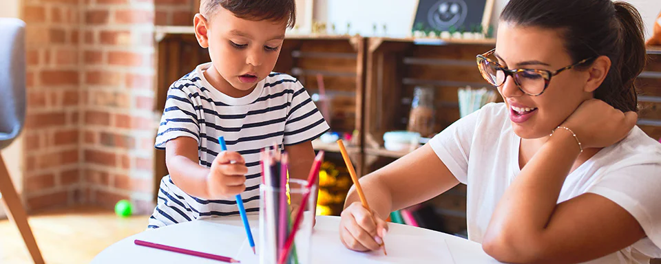 Toddler using colored pencils and interacting with smiling teacher