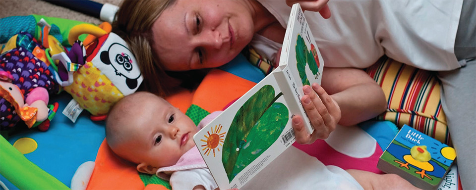 Mother laying on floor next to her baby while reading a book