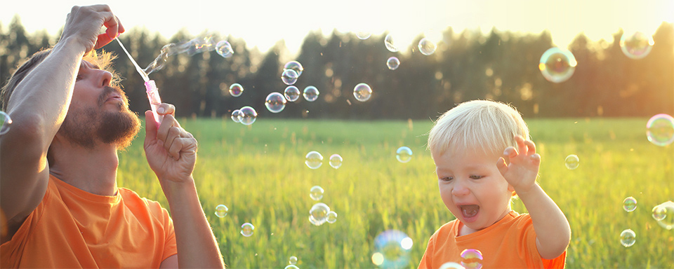 Child watches with delight as father blows bubbles in an open green field