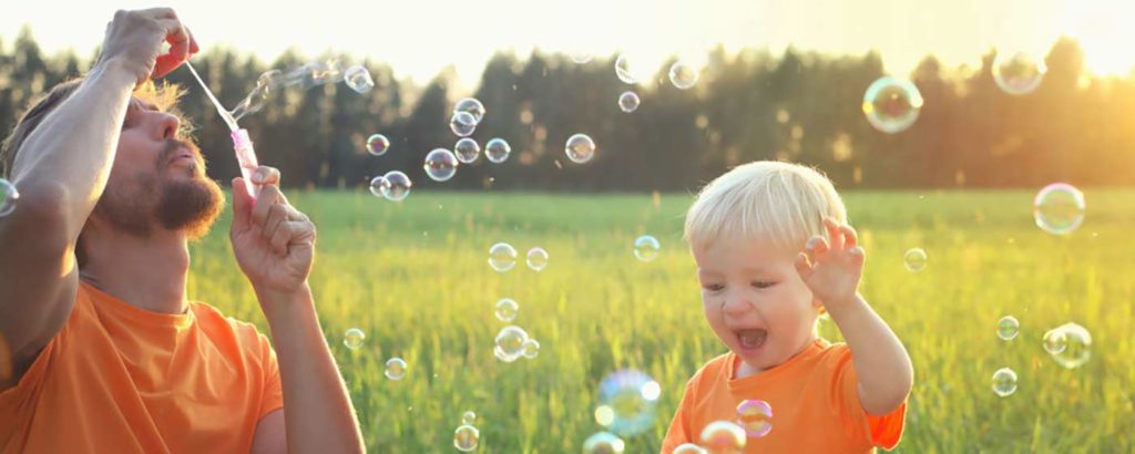 Child watches with delight as father blows bubbles in an open green field