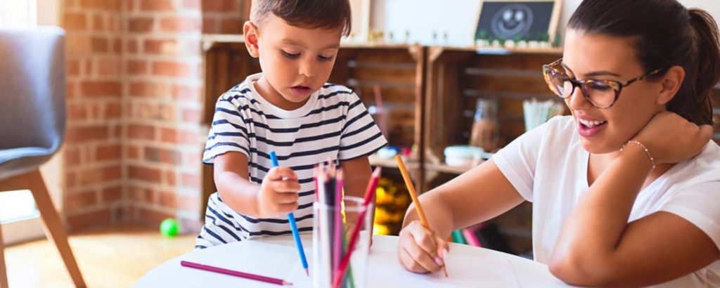 Toddler using colored pencils and interacting with smiling teacher